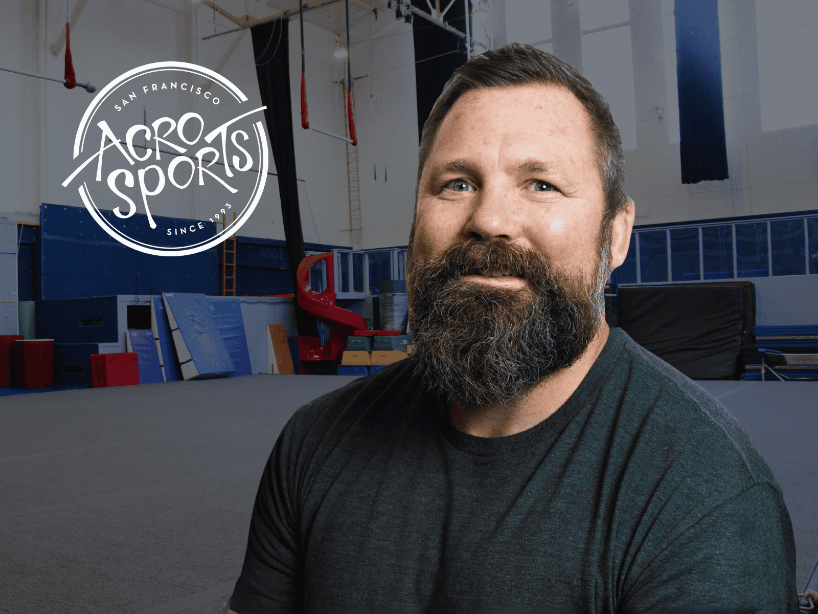 Johnny Bradford smiling inside the AcroSports San Francisco facility next to the AcroSports logo, celebrating his appointment as Chief Programs Officer.
